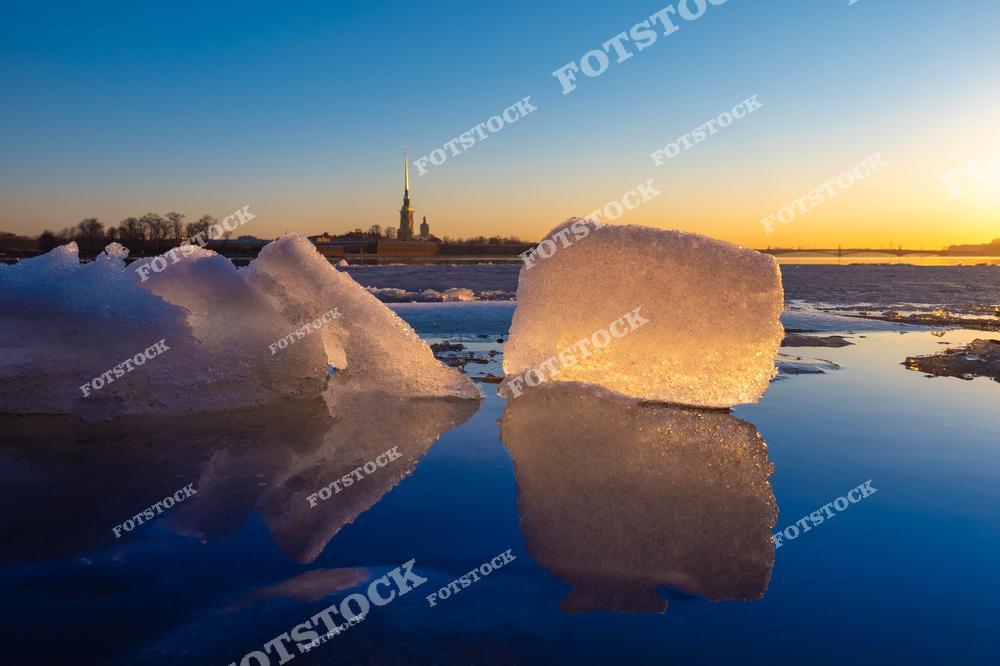 На переднем плане фотографии запечатлены крупные куски льда на замерзшей поверхности Невы, отражающие закатное небо. На заднем плане видна Петропавловская крепость с её величественным шпилем. Золотистый свет заката создает волшебную атмосферу, подчеркивая красоту зимнего Санкт-Петербурга. Это место притягивает своей природной и исторической гармонией.